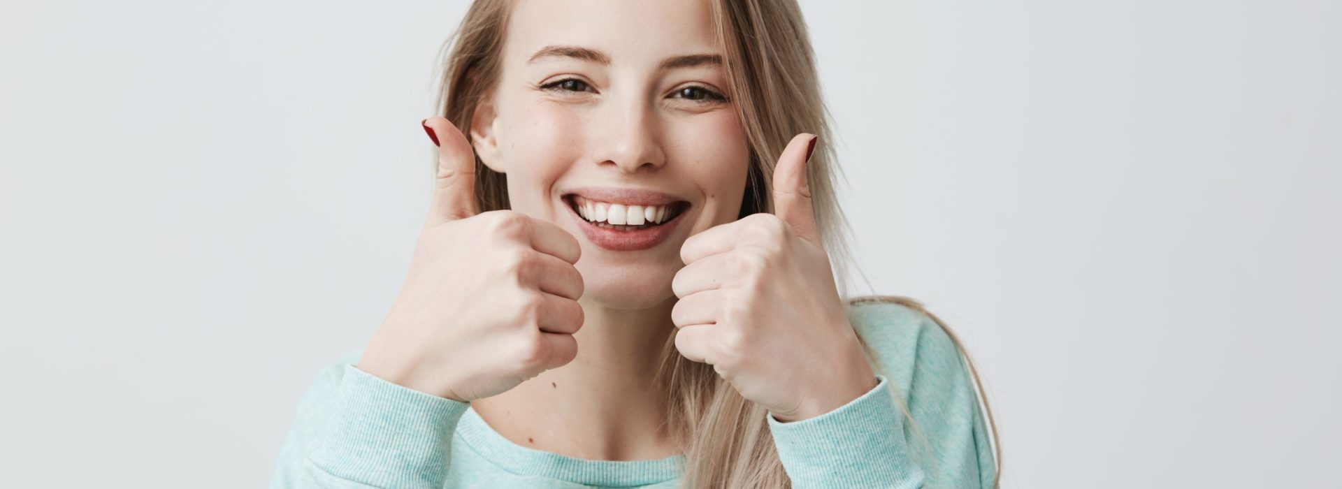 Portrait of positive blonde female student or customer with broad smile, looking at the camera with happy expression, showing thumbs-up with both hands, achieving goals. Body language