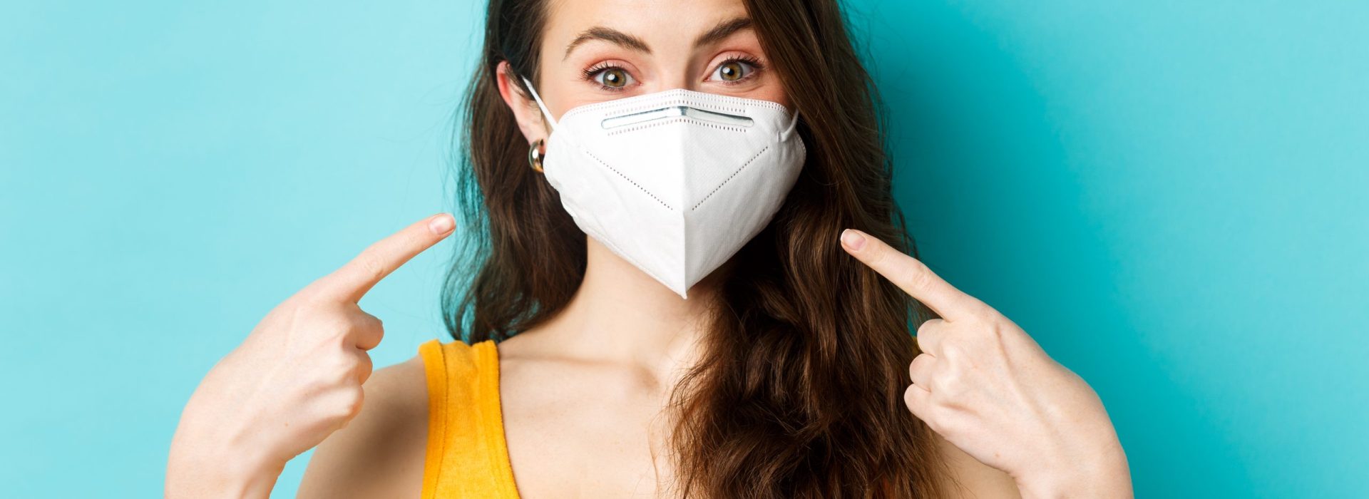 Covid-19, coronavirus and social distancing. Young woman in respirator pointing at her face, asking to use face masks during pandemic, standing against blue background.